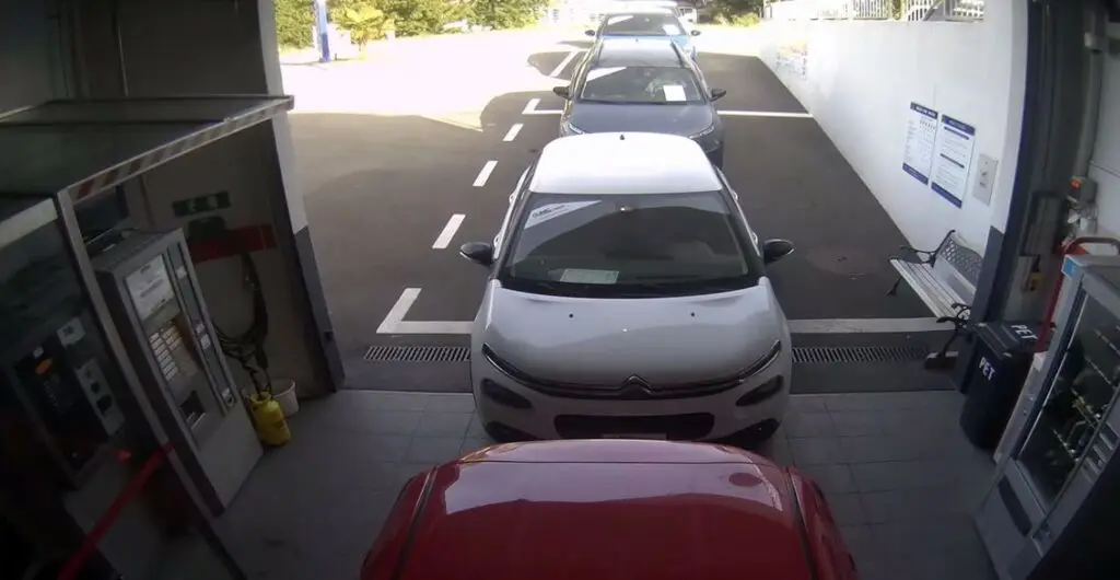 Une file de véhicules attend patiemment dans une voie unique d’un centre de contrôle ou de maintenance automobile. À l’avant, une voiture rouge est suivie d’une Citroën blanche et d’autres véhicules. Sur la gauche, des équipements techniques sont visibles, probablement pour le diagnostic ou le lavage. À droite, un banc, un distributeur automatique et une poubelle PET renforcent l’aspect fonctionnel et accueillant du lieu. L’environnement est propre, bien éclairé, et structuré pour un flux efficace.